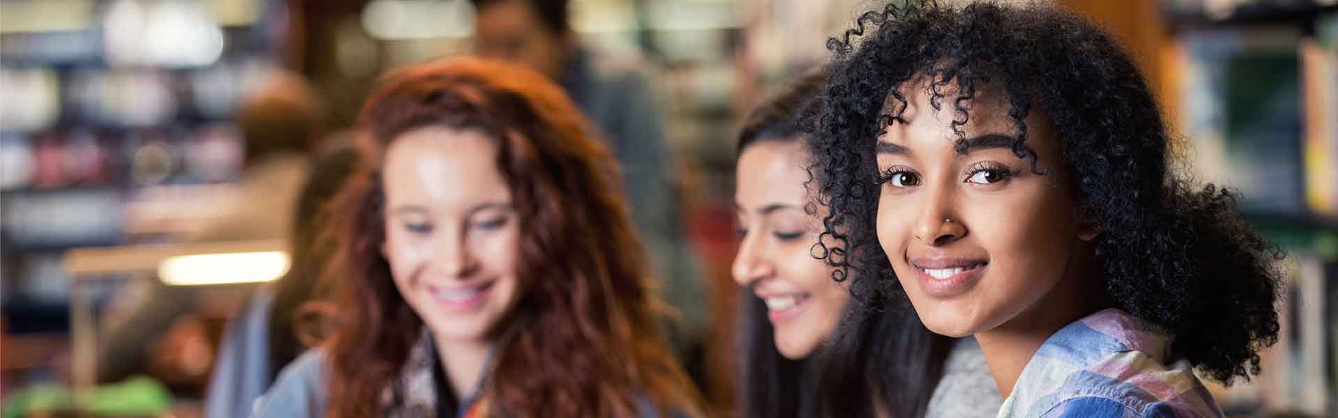 Three girls working at laptop in a library. Girl in foreground looks at camera.